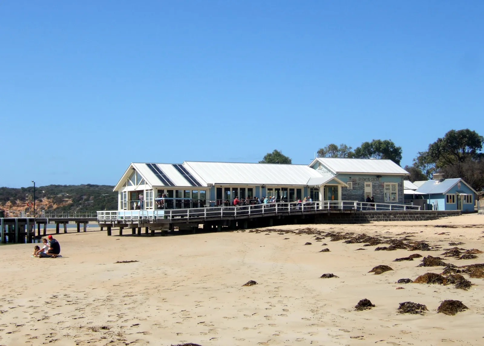 The restaurant on the timber jetty at Barwon Heads, with the small blue boatshed beside it and seaweed on the sand in front