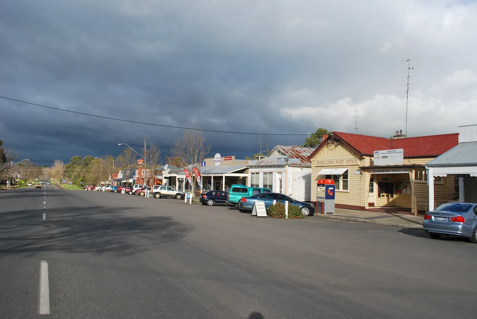 Birregurra's wide main street under storm clouds, with the weatherboard post office and a row of shops