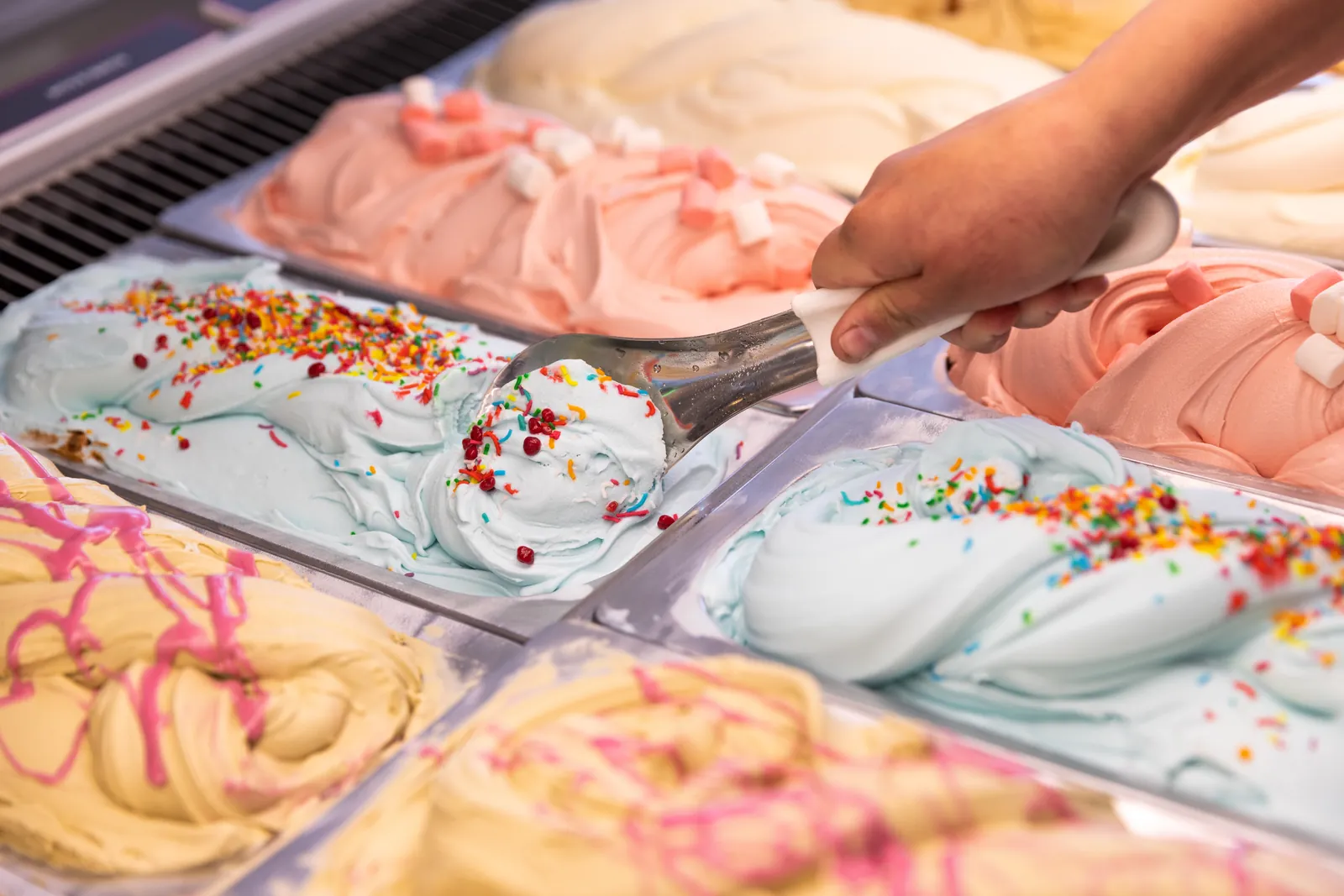 Scooping blue gelato topped with sprinkles from a cabinet of pastel flavours