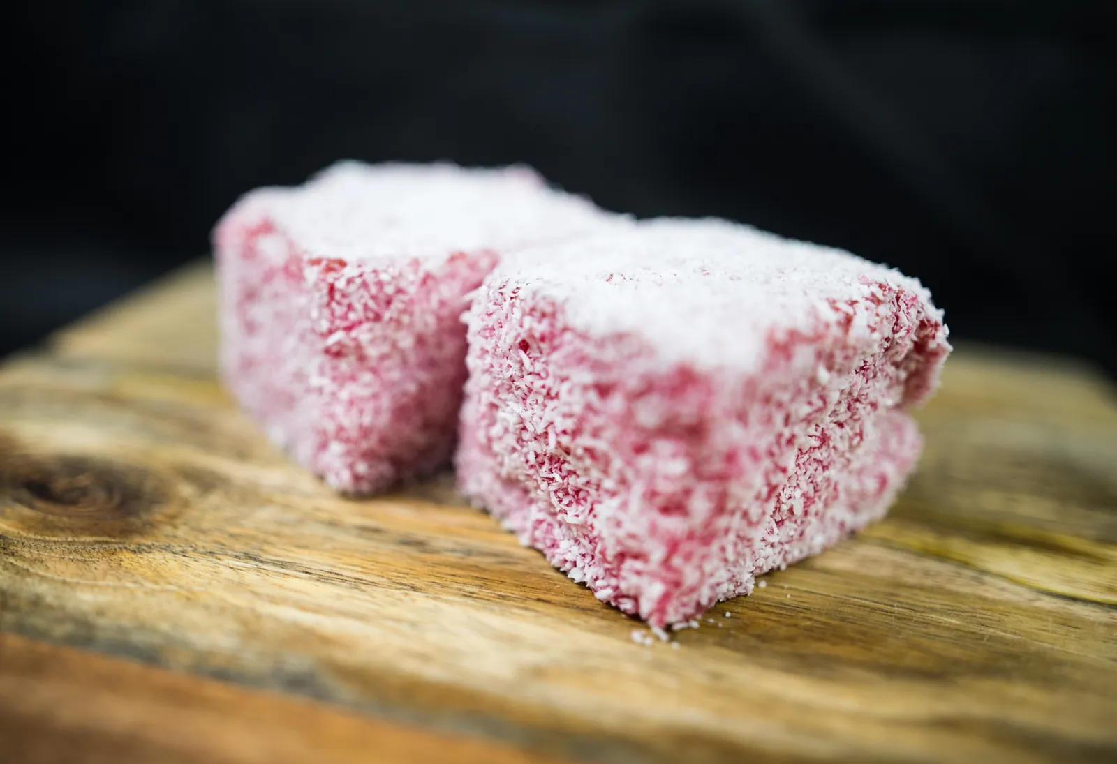 Two raspberry lamingtons rolled in coconut, on a timber board
