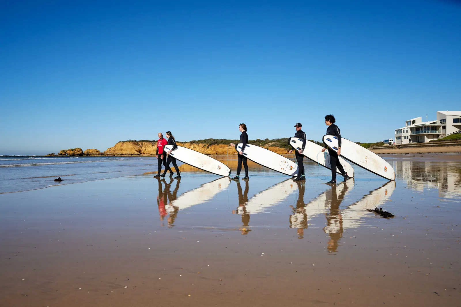 Four surfers carrying boards across the wet sand at Torquay during a surf lesson