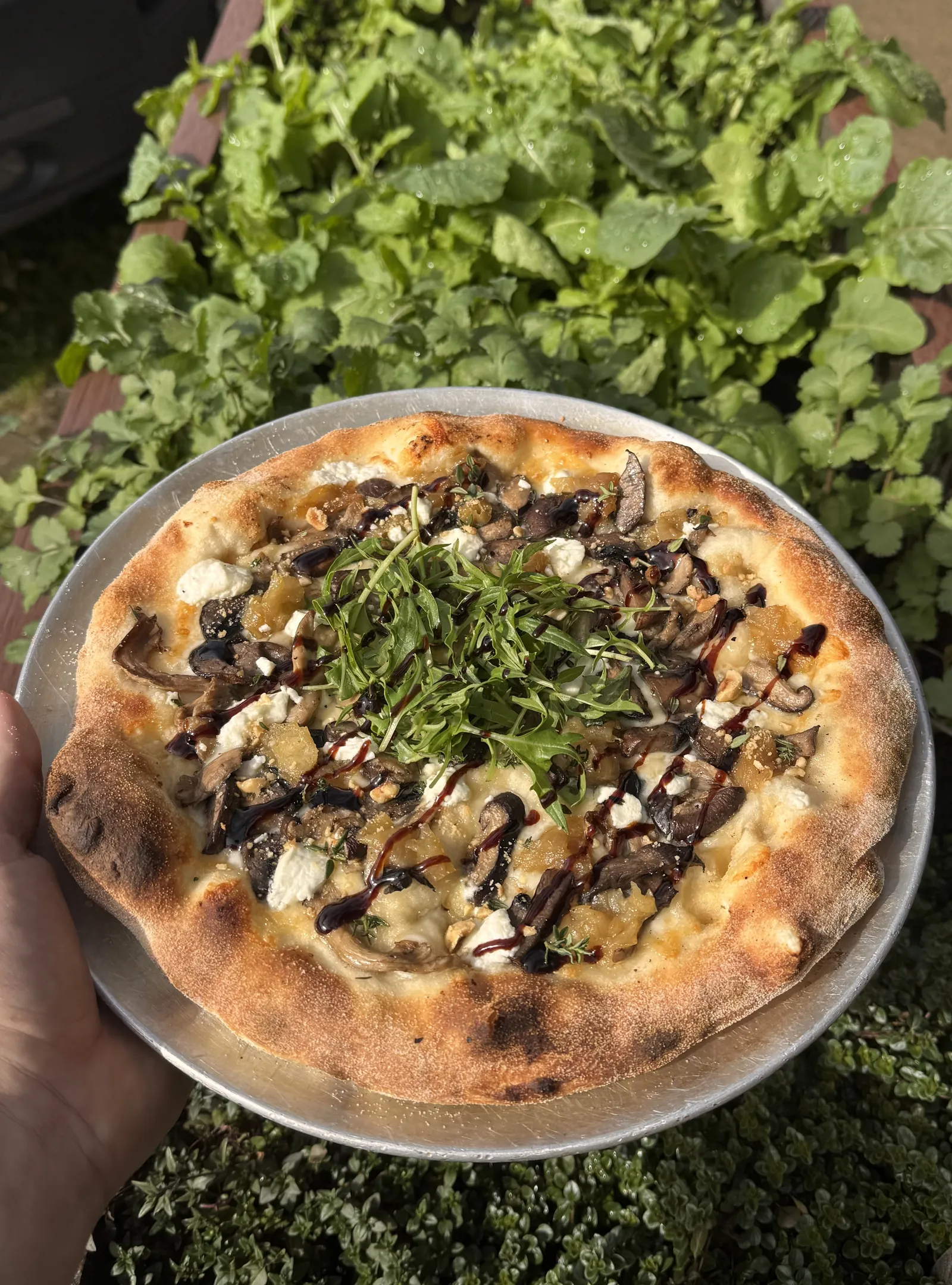 A wood-fired mushroom pizza topped with fresh rocket and a balsamic drizzle, held up over the garden