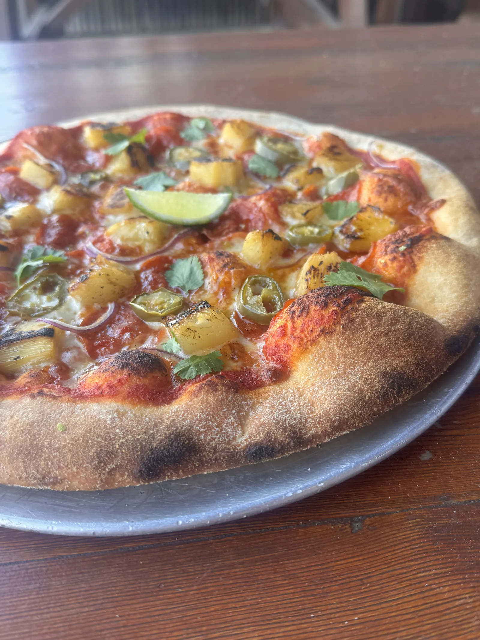 A wood-fired pizza with pepperoni, pineapple, jalapenos, coriander and a wedge of lime on a wooden table