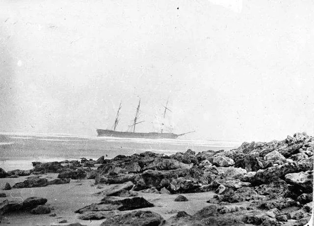 A black and white photograph of the Joseph H. Scammell aground on a reef, seen from a rocky beach