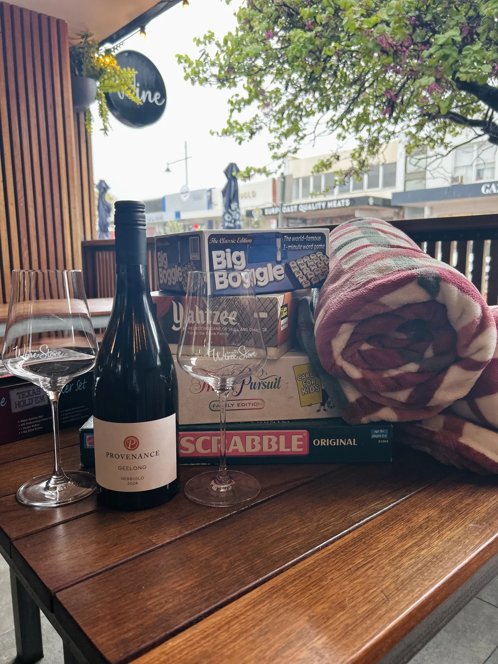 A bottle of red, glasses, board games and a rug on a table out the front of Torquay Wine Store