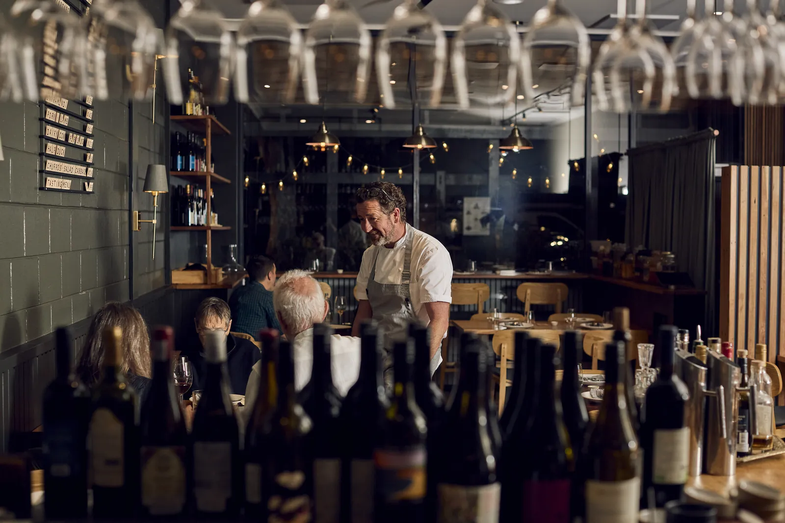 A chef serving diners at Samesyn, seen past a row of wine bottles