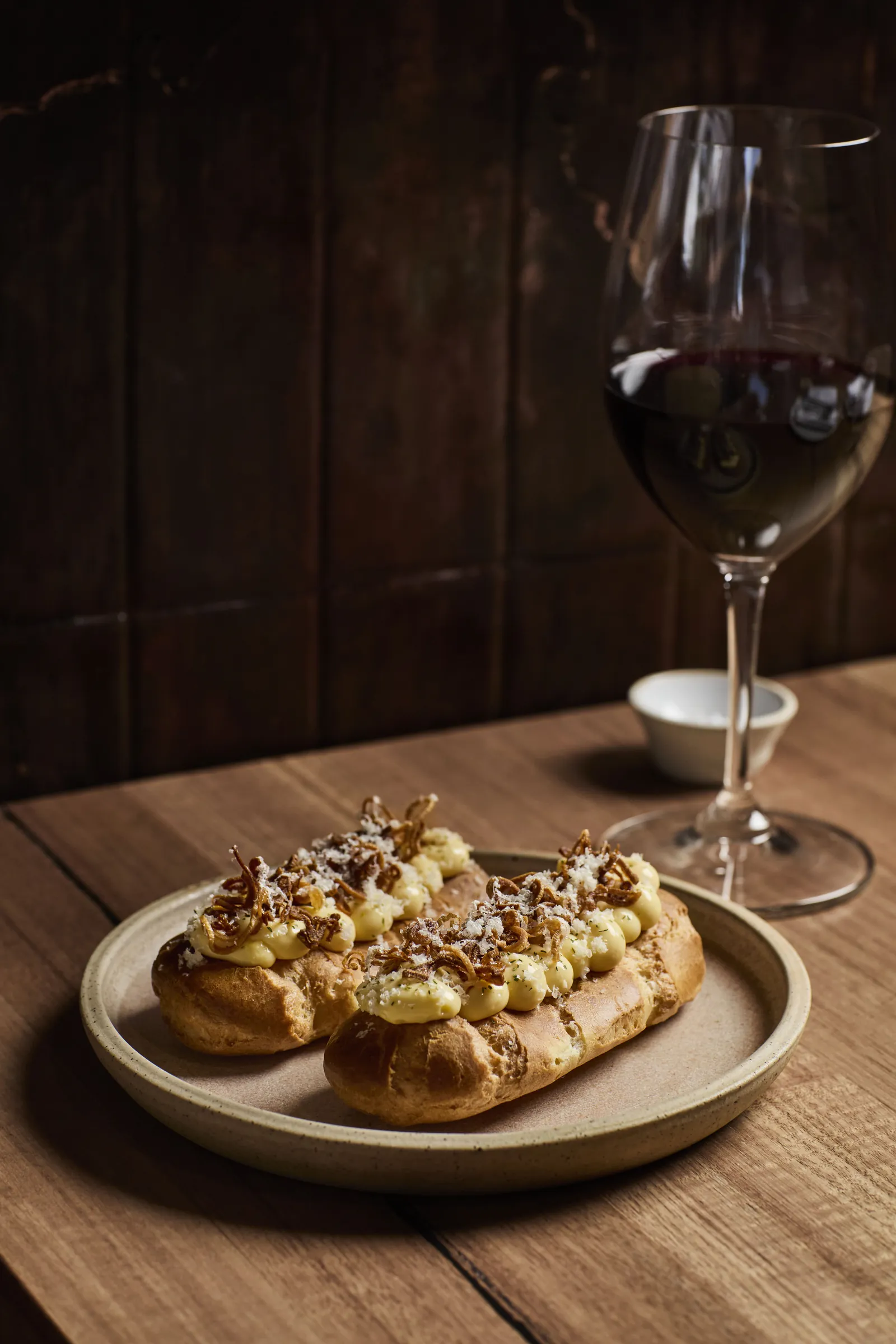 Two eclairs topped with nuts on a ceramic plate, beside a glass of red wine