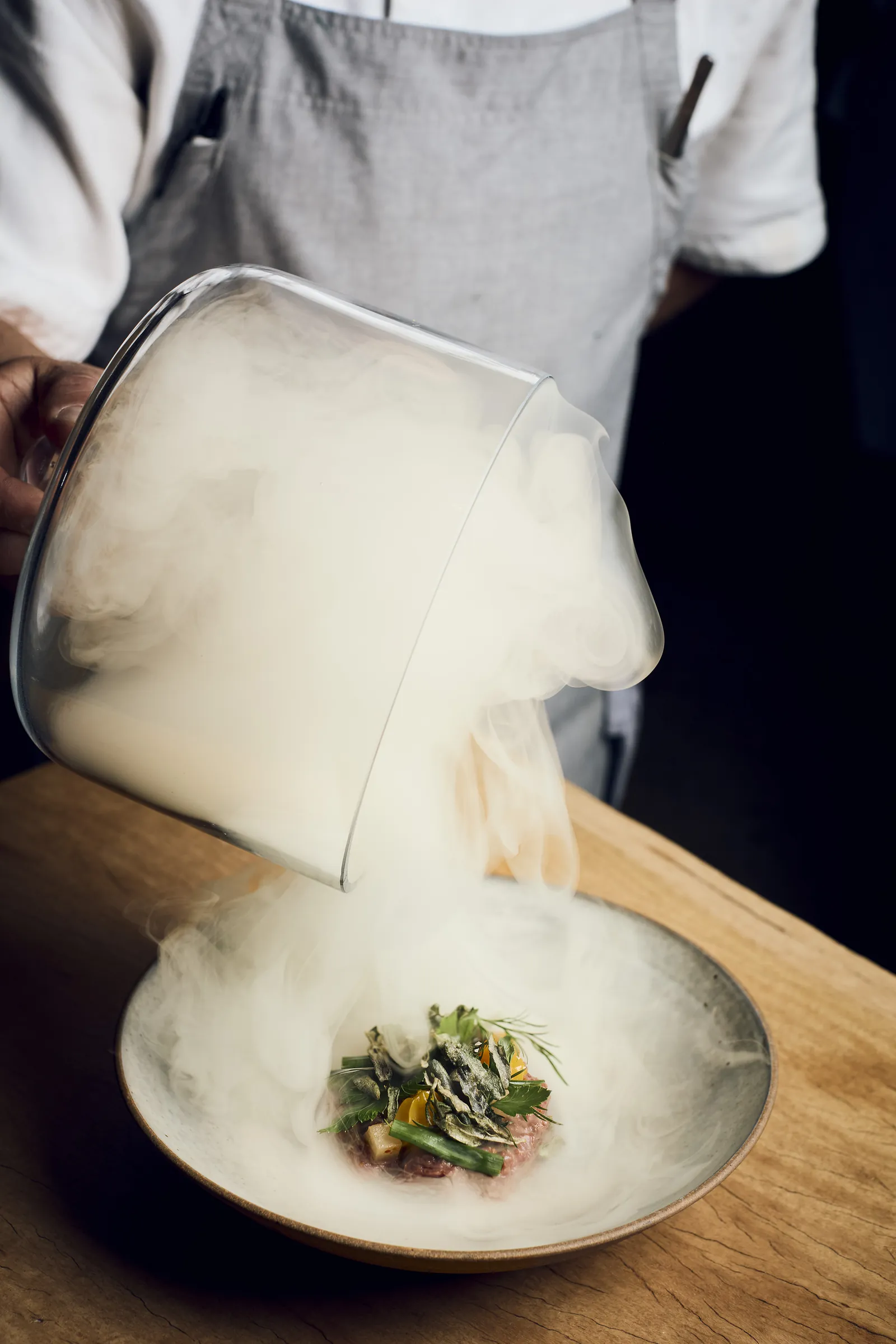 A chef lifting a glass cloche as smoke spills over a small plated dish